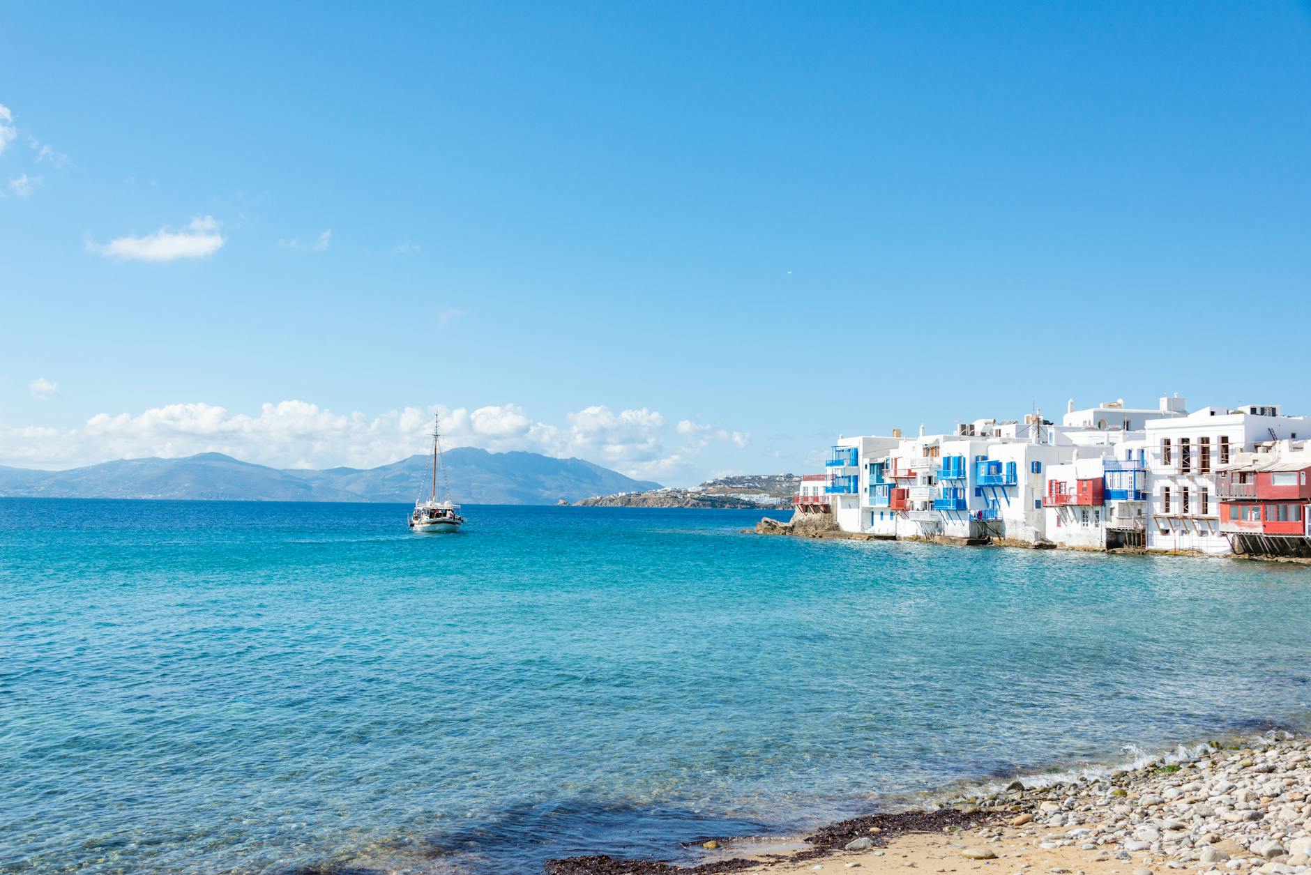 A stunning view of Mykonos with iconic white buildings and a sailboat on the turquoise water.